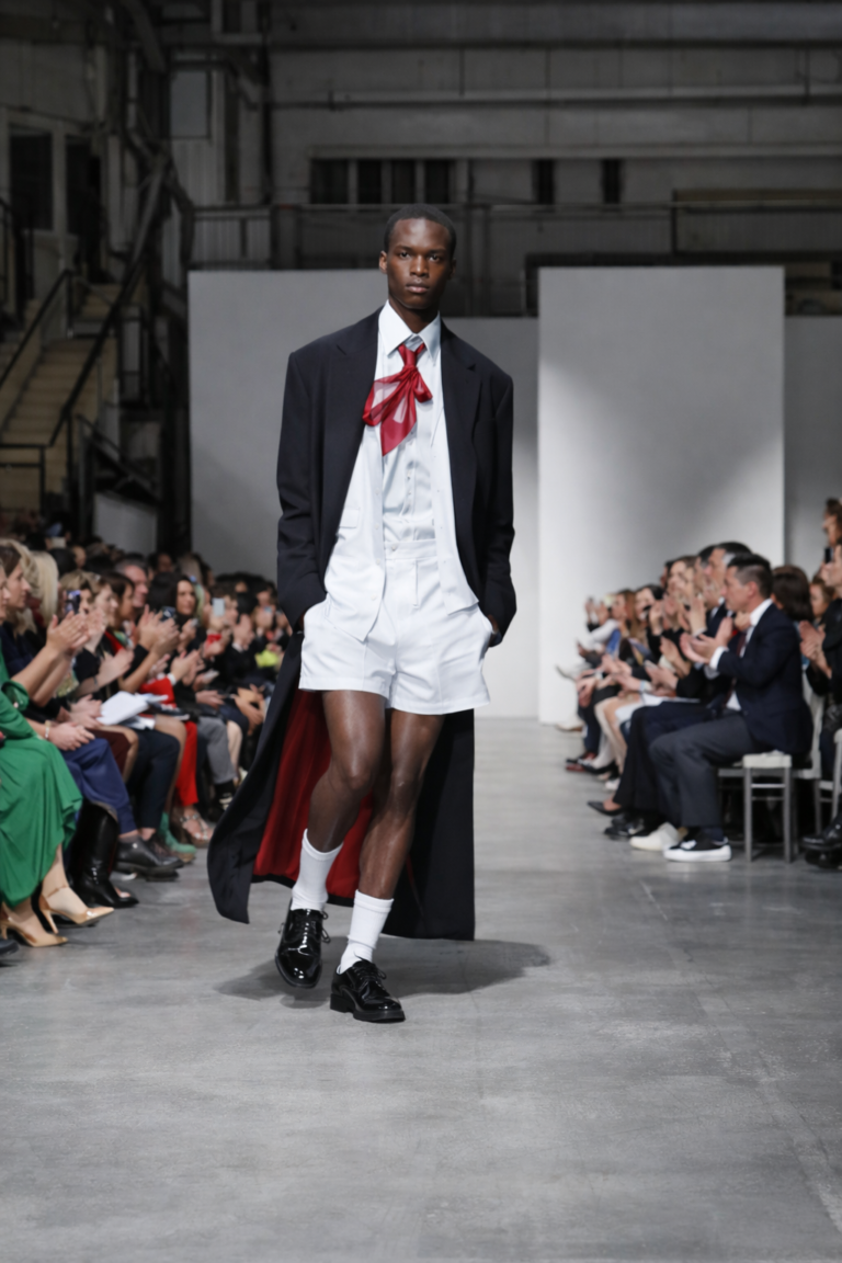 AI-generated image of a male model walking the runway in avant-garde menswear with a long black coat, white tailored shorts, and red ribbon tie