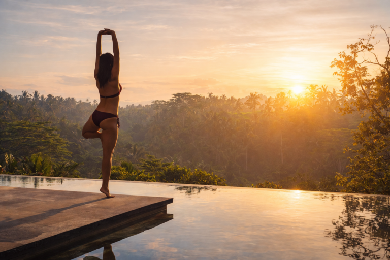 woman practicing yoga at sunrise on infinity pool terrace overlooking tropical forest landscape