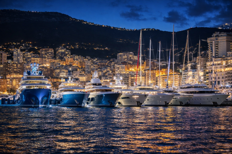 AI-generated image of luxury superyachts moored in Monaco’s harbor at night with the illuminated Monte Carlo skyline reflecting across the Mediterranean water.