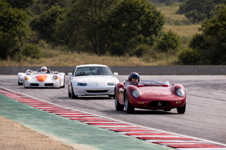 AI-generated image of classic vintage sports cars racing through a scenic motorsport circuit surrounded by trees and countryside.