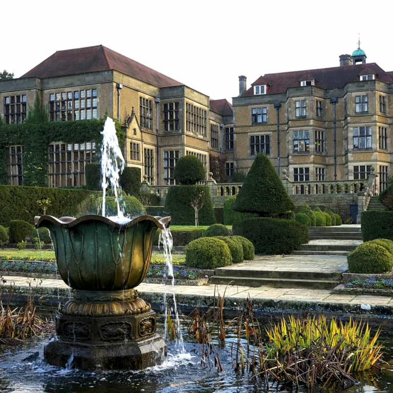 Historic English manor house surrounded by manicured formal gardens and a decorative fountain in the foreground